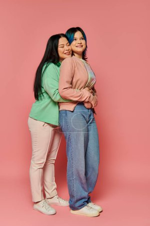 Asiatische Mutter und Teenagertochter in lässiger Kleidung stehen gemeinsam vor einem leuchtend rosa Hintergrund und strahlen Verbundenheit und Einheit aus.