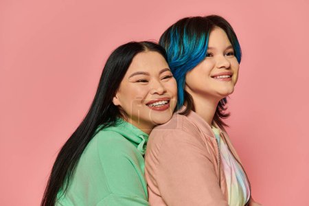 Téléchargez les photos : Mère asiatique et fille adolescente en tenue décontractée, toutes deux aux cheveux bleus, s'embrassant joyeusement en studio sur fond rose. - en image libre de droit