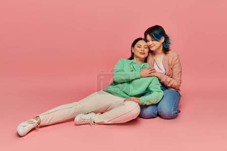 Asiatische Mutter und Teenager-Tochter in Freizeitkleidung sitzen eng beieinander auf rosa Hintergrund.