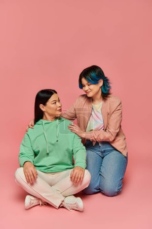 An Asian mother and her teenage daughter sitting next to each other in a studio, both wearing casual clothing, on a pink background.