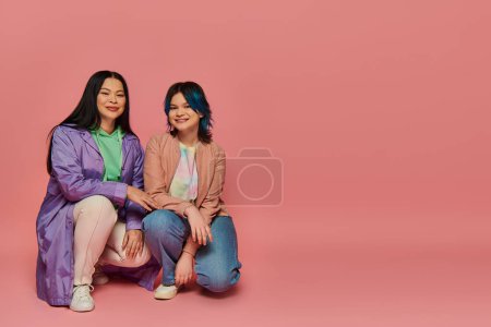 Photo for Asian mother and teenage daughter in casual wear sitting closely next to each other on a vibrant pink background. - Royalty Free Image