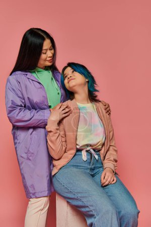 Téléchargez les photos : Two women, an Asian mother and her teenage daughter, stand together in casual wear against a vibrant pink background. - en image libre de droit