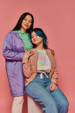 An Asian mother and her teenage daughter, dressed in casual wear, posing together in a studio against a pink background.