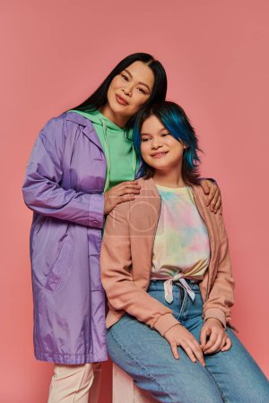 Foto de Una madre asiática y su hija adolescente, se sientan lado a lado en atuendo casual contra un fondo rosa vibrante. - Imagen libre de derechos