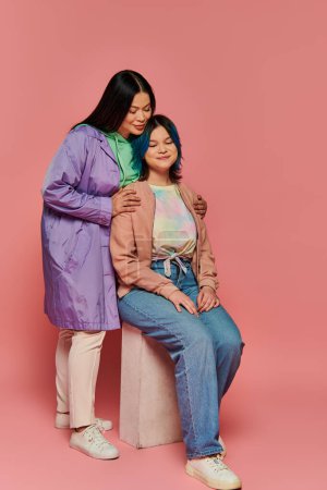 An Asian mother and her teenage daughter, both in casual wear, sit together on a bench against a vibrant pink background.