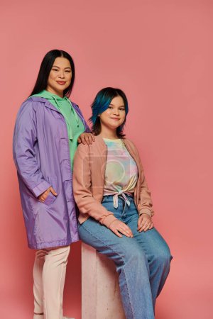 Asian mother and her teenage daughter standing together in casual wear, showcasing bond on pink studio background.