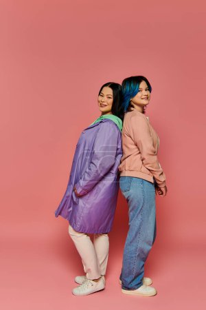 Téléchargez les photos : An Asian mother and her teenage daughter stand together in a casual pose against a vibrant pink background. - en image libre de droit