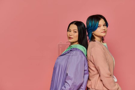 A stylish Asian mother and her teenage daughter, both with vibrant blue hair, pose together on a pink studio background.