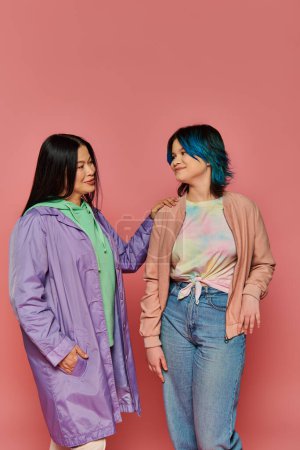Asian mother and teenage daughter in casual wear stand together in front of a vibrant pink wall.