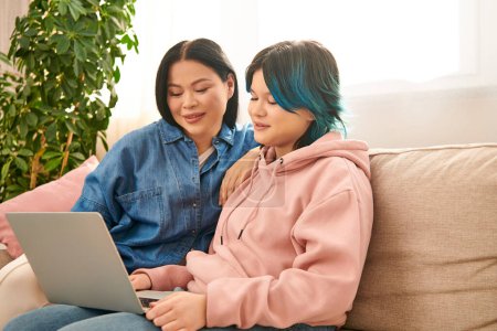 an Asian mother and her teenage daughter, sit on a couch and focus on a laptop screen in a cozy living room.