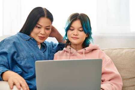 Asian mother and teenage daughter in casual wear sitting on a couch, engrossed in a laptop together.
