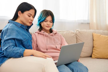 Asian mother and teenage daughter in casual wear sitting on a couch, engrossed in a laptop screen.