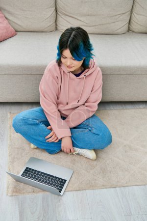Una chica con el pelo azul se sienta al lado de un portátil en el suelo, profundamente en el pensamiento