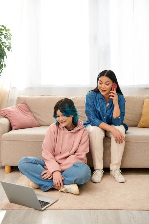 Asian mother and daughter in casual wear, seated on a couch, engaged in separate digital world