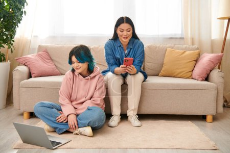 Asian mother and her teenage daughter, wearing casual attire, are sitting on a couch and focused on a cell phone screen.