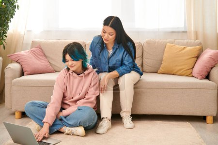 Asian mother and her teenage daughter sitting on a couch, focused on a laptop screen in a cozy home setting.