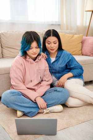 An Asian mother and her teenage daughter, in casual attire, sit on the floor together, engrossed in using a laptop.