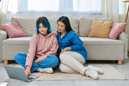 A mother and her teenage daughter, in casual attire, sitting on the floor, fully engaged with a laptop screen.