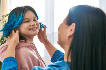 An Asian mother with blue hair gently combing her teenage daughters hair in a casual home setting.