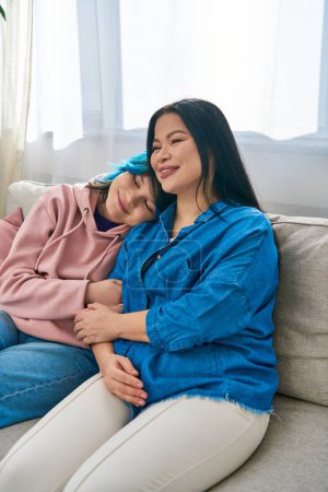 Foto de Una madre asiática y su hija adolescente en ropa casual se sientan juntas encima de un sofá, compartiendo un momento de cercanía. - Imagen libre de derechos