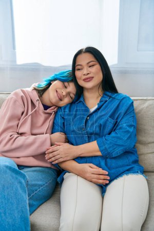 Téléchargez les photos : Une mère asiatique et sa fille adolescente en tenue décontractée, assise sur un canapé, partageant du temps de qualité ensemble à la maison. - en image libre de droit