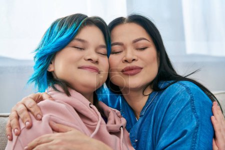 Mother and daughter, both Asian, share a comforting hug on a cozy couch, displaying love and family bonding.