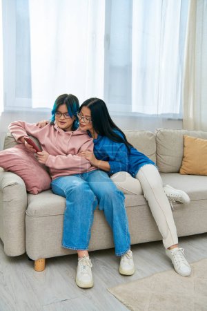 An Asian mother and her teenage daughter, in casual wear, sit together on a cozy couch in their living room.