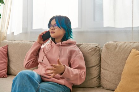 An Asian girl with blue hair sit on a couch, talking on a cell phone together.