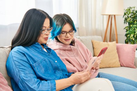 Una madre asiática y su hija adolescente se sientan en un sofá, absortos en un teléfono celular.
