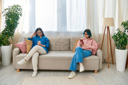 Photo for An Asian mother and her teenage daughter sit on a couch in a living room, spending time together in casual wear. - Royalty Free Image