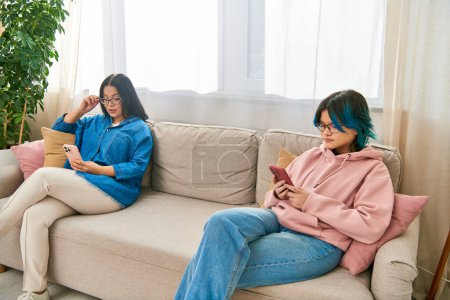 Téléchargez les photos : Mère asiatique et la fille adolescente en tenue décontractée assis sur un canapé, absorbé dans leurs téléphones, passer du temps ensemble à la maison. - en image libre de droit