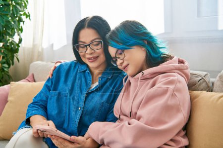 Asian mother and daughter, in casual attire, sit on a couch, focused on a cellphone screen, bonding and spending quality time together.