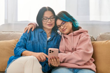 Foto de Una madre y una hija adolescente, ambas asiáticas, en ropa casual, sentadas cómodamente encima de un sofá juntas. - Imagen libre de derechos