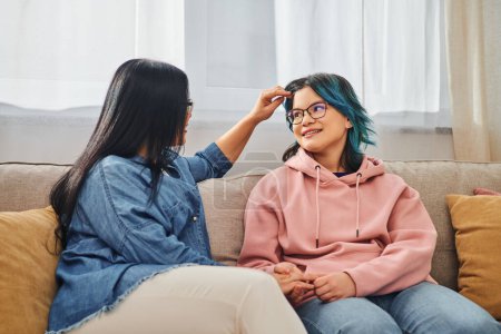 An Asian mother sitting on a couch, tenderly stroking her teenage daughter hair in a cozy home setting.