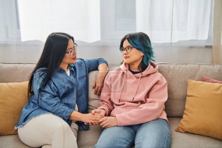 Foto de Una madre asiática y su hija adolescente en ropa casual entablan una conversación profunda mientras se sientan en un sofá acogedor. - Imagen libre de derechos