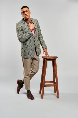 Fashionable man stands by a stool in a plaid blazer.
