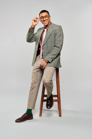 Man in plaid blazer and tie sitting on stool.