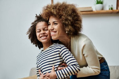 A happy African American mother and daughter embrace affectionately on a cozy couch at home.
