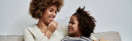 Afroamerikanische Mutter und Tochter teilen sich ein Lächeln, während sie auf einer gemütlichen Couch sitzen und die gemeinsame Zeit zu Hause genießen.