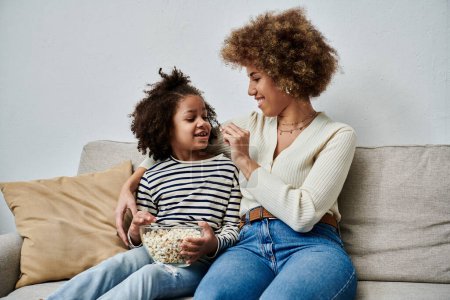Photo for An African American mother and daughter happily sit on a couch, enjoying popcorn together. - Royalty Free Image