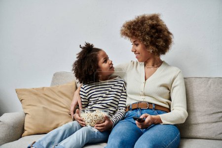 Feliz madre afroamericana y su hija compartiendo un momento en el sofá mientras ven la televisión juntos.