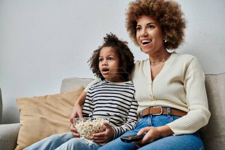 Happy African American mother and daughter sit together on a couch, watching TV with smiles on their faces.