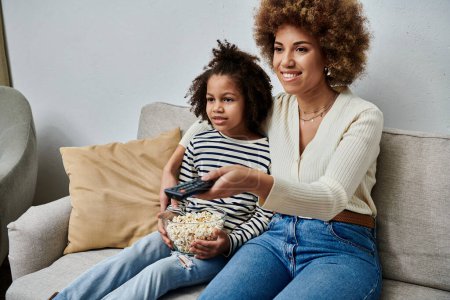 Amar a la madre y a la hija afroamericanas felizmente se sientan en un sofá, absortos en ver la televisión juntos.