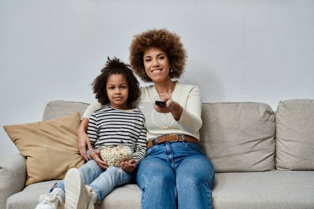 A joyful African American mother and her daughter of African descent sitting on a couch, engrossed in watching television.