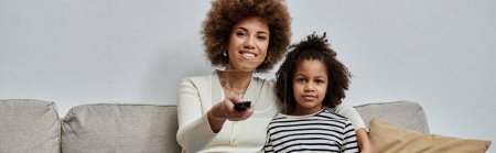 Téléchargez les photos : A happy African American mother and daughter sitting on a couch, holding a remote control, enjoying quality time together at home. - en image libre de droit