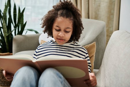 Foto de Un niño afroamericano se sienta cómodamente en un sofá, lee un libro - Imagen libre de derechos