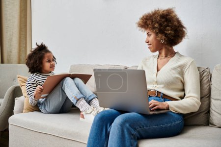 Happy African American mother and daughter sitting on a couch, using a laptop together at home.