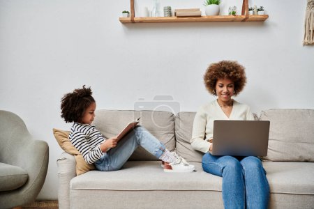 An African American mother and daughter happily seated on a couch, engrossed in using a laptop together.