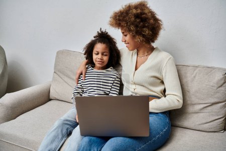 Téléchargez les photos : A happy African American mother and daughter sitting on a couch, engrossed in using a laptop together. - en image libre de droit