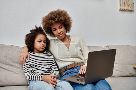Photo for A happy African American mother and daughter sit on a couch using a laptop together. - Royalty Free Image